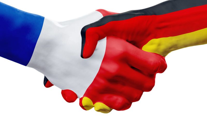 Zwei Hände reichen sich zum Handschlag, eine ist in den Farben der französischen Flagge, die andere in den Farben der deutschen Flagge eingefärbt. Das Bild symbolisiert die enge Partnerschaft zwischen Frankreich und Deutschland.
