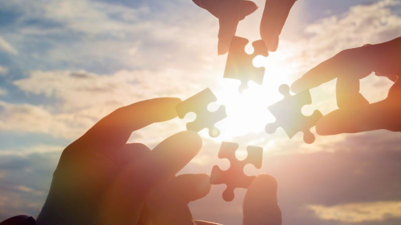 Vier Hände halten vier zueinander passende Puzzle-Teile vor einen stimmungsvoll beleuchteten Himmel. Ein Symbolbild für Zusammenarbeit.