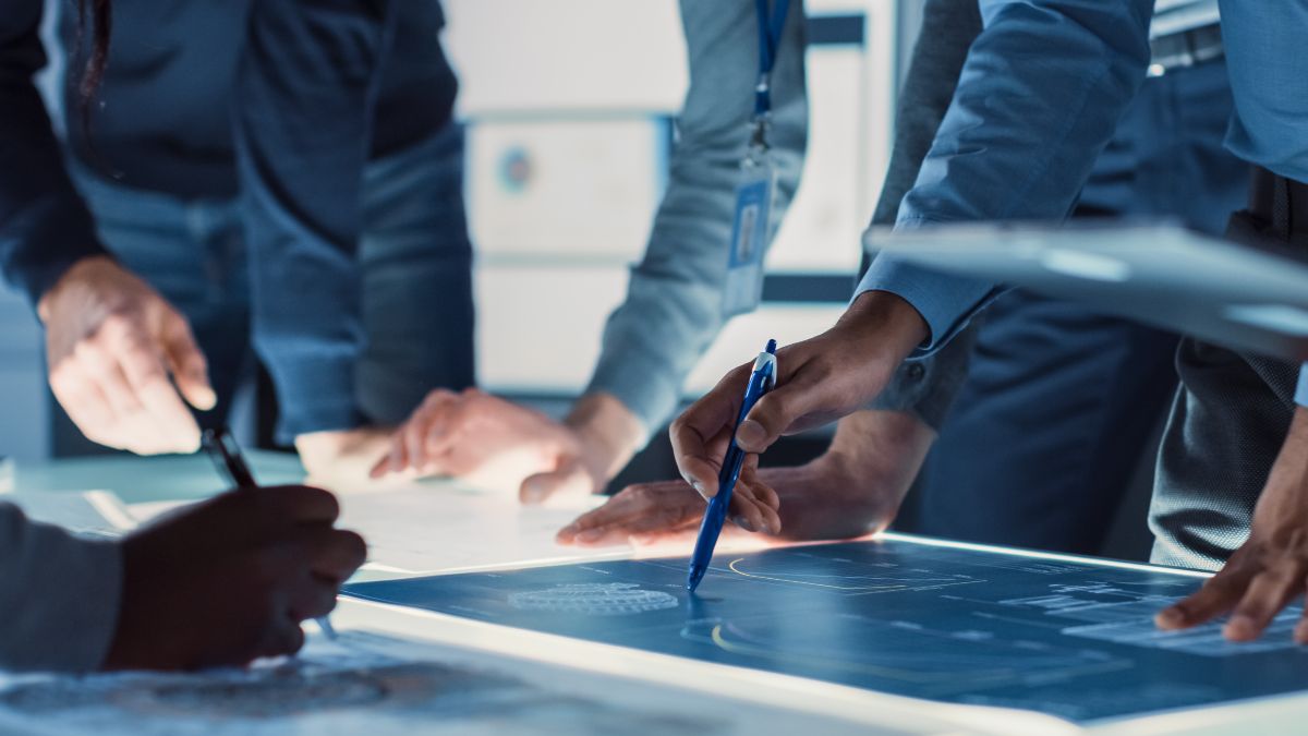 Mehrere Personen arbeiten gemeinsam an einem beleuchteten Tisch mit digitalen Plänen, zeigen darauf und markieren Inhalte. Ein Symbol für Teamarbeit und Innovation.